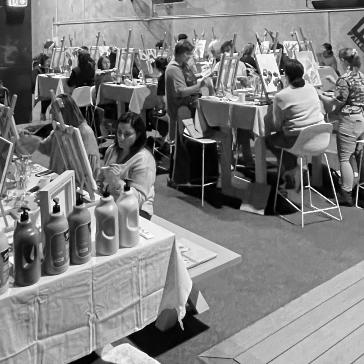 Black and white photo of a large indoor space with people sitting at tables, painting in a creative workshop, in warehouse space.