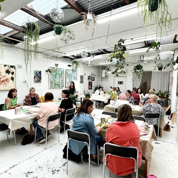 Group of people in a painting workshop, sitting at tables in a room with plants and artwork on the walls.