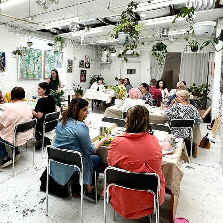 Group of people in a painting workshop, sitting at tables in a room with plants and artwork on the walls.