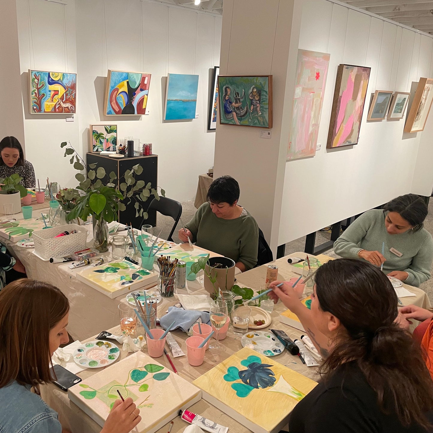Team of people painting at a table in an art studio and gallery with various artworks on the walls. The team are painting botanicals together.