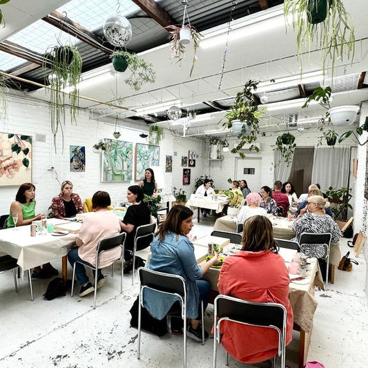 Group of people in a painting workshop, sitting at tables in a room with plants and artwork on the walls.