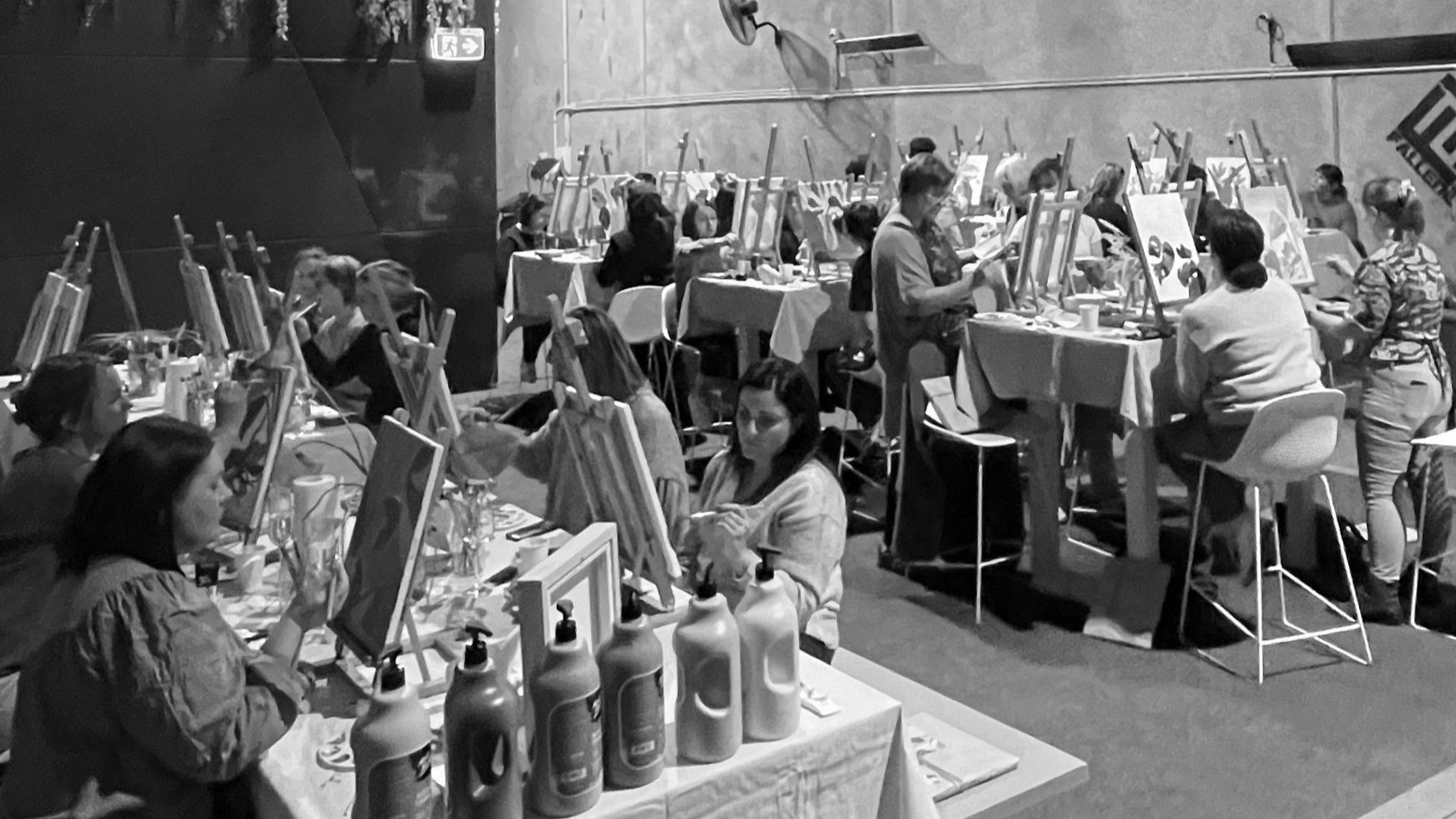 Black and white photo of a large indoor space with people painting in a workshop sitting at tables, string lights hanging from the ceiling.