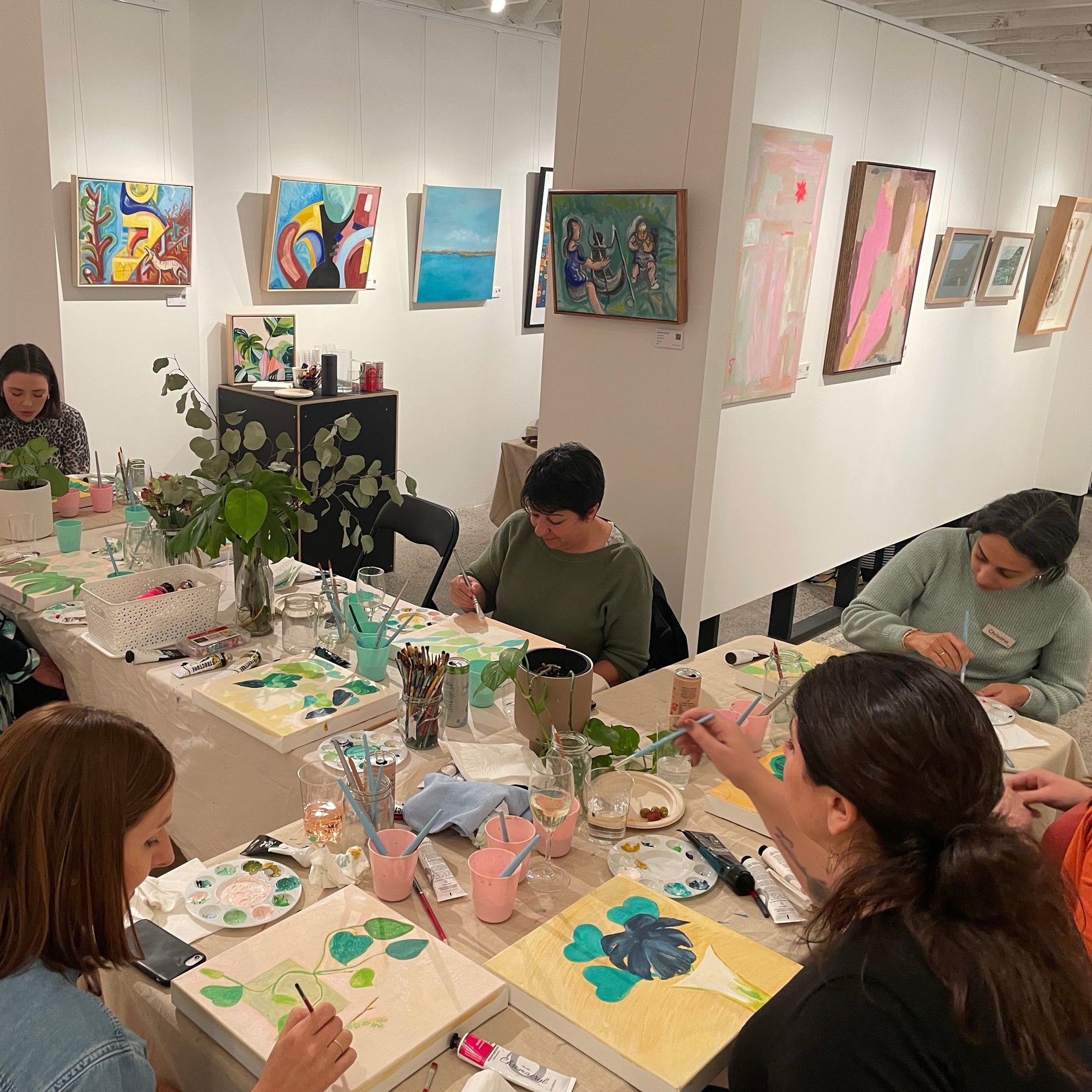 Team of people painting at a table in an art studio and gallery with various artworks on the walls. The team are painting botanicals together. 